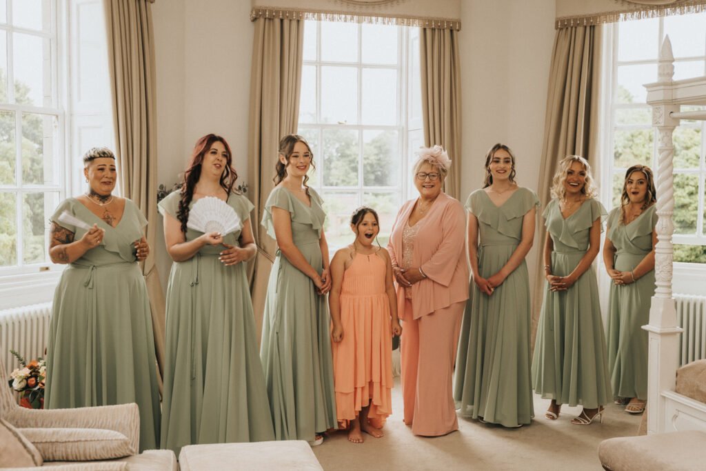 Seven women in sage green dresses and a girl in a peach dress stand smiling in a bright room with large windows and beige curtains. One woman in the centre wears a peach outfit and headpiece. The atmosphere is joyful and celebratory. © Aimee Lince Photography, Lincolnshire photographer