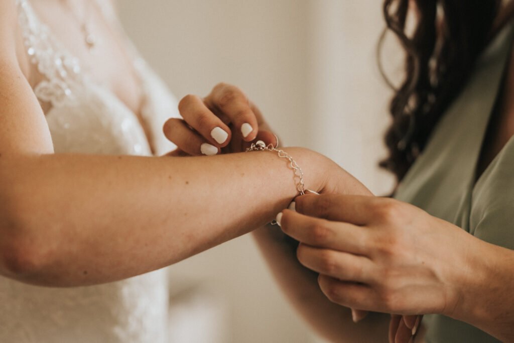 Close-up of one woman fastening a delicate silver bracelet on another woman’s wrist. The woman receiving help wears a white lace dress, suggesting she is a bride. Both women have manicured nails and soft lighting creates a warm, intimate feel. © Aimee Lince Photography, Lincolnshire photographer