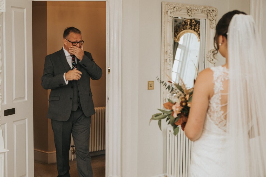 A man in a grey suit stands in a doorway, covering his mouth in emotional surprise. A bride in a white lace dress and veil, holding a bouquet, faces him with her back to the camera. The scene is indoors with a large ornate mirror on the wall. © Aimee Lince Photography, Lincolnshire photographer