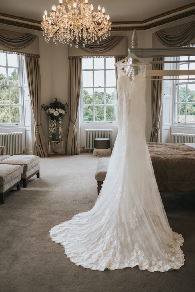 A lace wedding dress on a mannequin stands in an elegant, softly lit room with tall windows, taupe curtains, a crystal chandelier, beige carpet, two benches, and a flower arrangement in the background. Natural light fills the space. © Aimee Lince Photography, Lincolnshire photographer