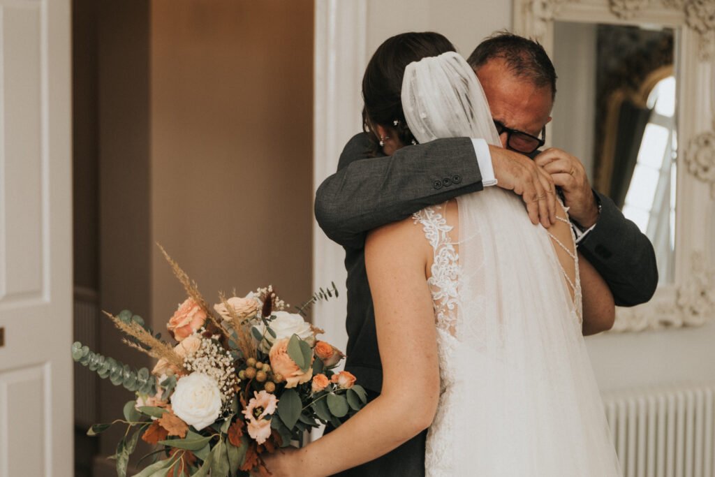 A bride in a white lace dress and veil hugs an older man in a suit, who appears emotional and wipes his eyes. She holds a bouquet of white and peach flowers with greenery. They stand indoors near a large ornate mirror and cream-coloured walls. © Aimee Lince Photography, Lincolnshire photographer