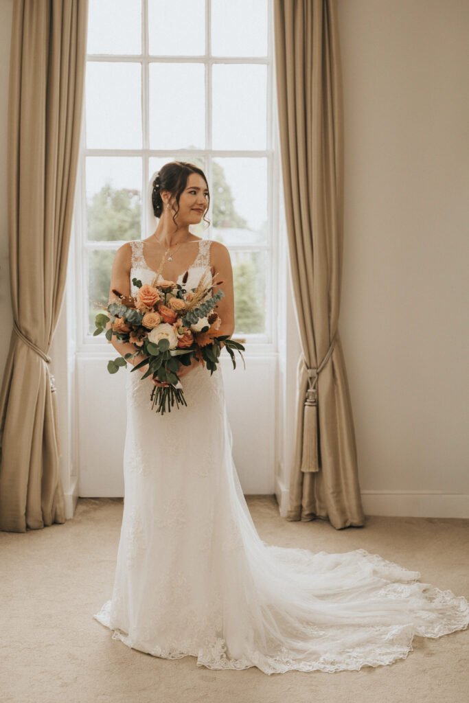 A bride in a white lace wedding dress stands indoors in front of tall windows with beige curtains. She holds a bouquet of orange, pink, and white flowers with greenery and gazes to the side, smiling softly. The room is bright and elegant. © Aimee Lince Photography, Lincolnshire photographer