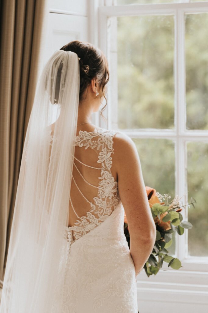 A bride in a lace wedding dress with a sheer back and delicate beadwork stands facing a window. She holds a bouquet of flowers and wears a long veil draped over her hair, with natural light streaming in from outside. © Aimee Lince Photography, Lincolnshire photographer