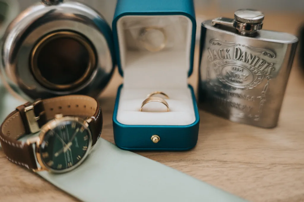 A close-up of a wooden surface displays a wristwatch with a brown strap, a turquoise ring box with two gold rings inside, a hip flask engraved with "Jack Daniel’s," and a round shiny metal object. The items suggest a wedding or special occasion. © Aimee Lince Photography, Lincolnshire photographer