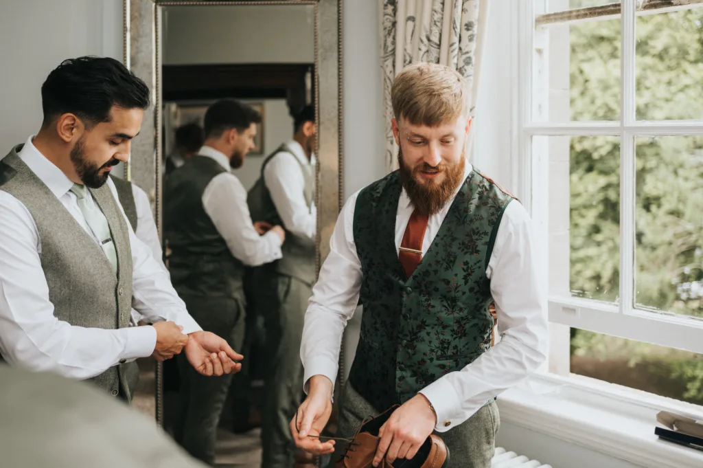 Two men in dress shirts and waistcoats stand near a window. One, with a beard, holds brown shoes and smiles, while the other adjusts his cuff. Both are reflected in a large mirror behind them. Sunlight streams through the window, illuminating the room. © Aimee Lince Photography, Lincolnshire photographer