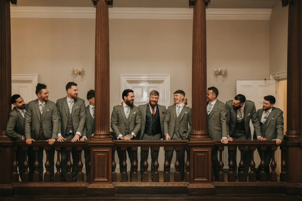 Eight men in grey suits and ties stand behind a wooden balcony railing in a formal room, smiling and talking. The man in the centre wears a darker suit and red tie, suggesting he is the groom, surrounded by groomsmen in matching attire. © Aimee Lince Photography, Lincolnshire photographer