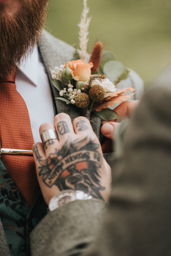 A close-up of a wedding photographer capturing a hand pinning a buttonhole of white and orange flowers onto a suit lapel, highlighting bold rings, tattoos, an orange tie, and tweed jacket with a patterned waistcoat. © Aimee Lince Photography, Lincolnshire photographer
