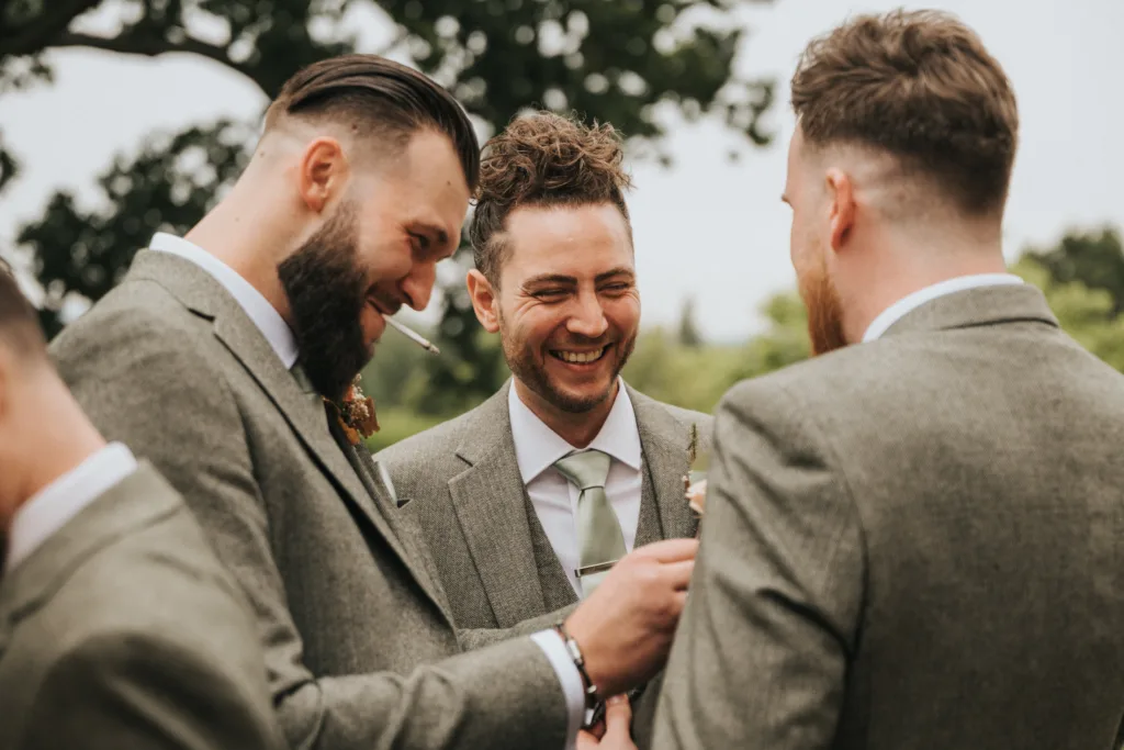 Three men in grey suits with buttonholes stand together outdoors, smiling and laughing. One is pinning a buttonhole on another’s jacket. They have groomed hair and beards, and trees are visible in the blurred background. © Aimee Lince Photography, Lincolnshire photographer