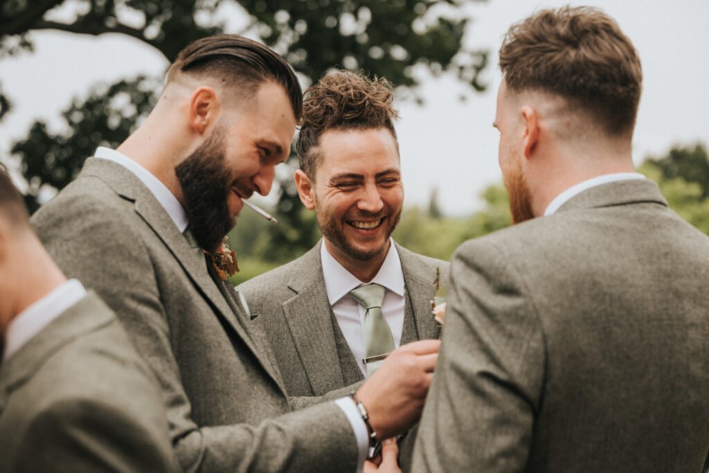 Three men in grey suits with buttonholes stand together outdoors, smiling and laughing. One is pinning a buttonhole on another’s jacket. They have groomed hair and beards, and trees are visible in the blurred background. © Aimee Lince Photography, Lincolnshire photographer
