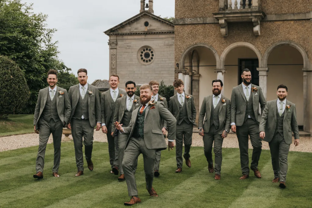 A smiling groom and eight groomsmen in matching grey suits walk across a lawn towards the camera. The groom is in front, looking joyful with outstretched arms. Behind them is a stone building with arches and greenery on the sides. © Aimee Lince Photography, Lincolnshire photographer