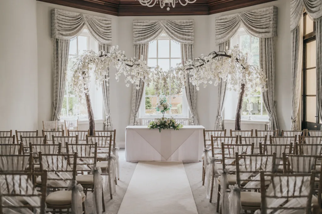Elegant indoor wedding venue with rows of wooden chairs draped in white fabric, facing a white aisle and a table decorated with flowers. Two arching trees with white blossoms frame the table, set in front of large windows with ornate curtains. © Aimee Lince Photography, Lincolnshire photographer