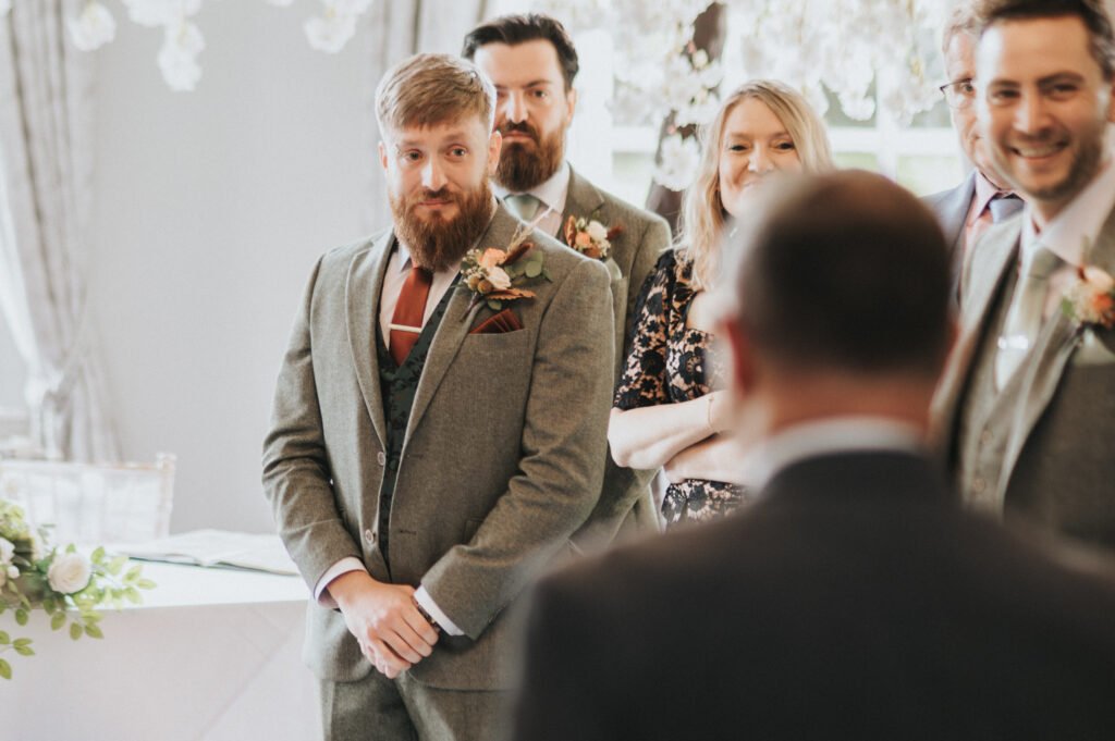 A bearded man in a grey suit with a buttonhole stands indoors, looking ahead and smiling slightly. Four people—two men and two women—stand nearby, dressed formally and smiling. White flowers and light curtains decorate the bright room. © Aimee Lince Photography, Lincolnshire photographer