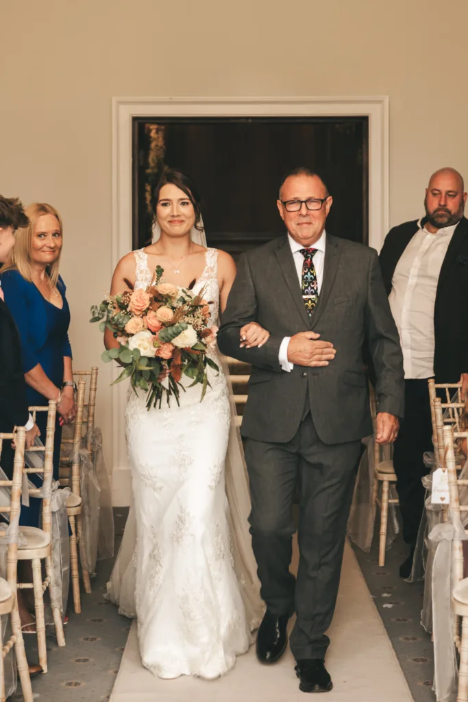 A bride in a white lace gown holding a bouquet walks down the aisle arm-in-arm with an older man in a grey suit. Guests seated on either side watch. The mood is joyful, and the room is softly lit with neutral decor and wooden chairs. © Aimee Lince Photography, Lincolnshire photographer
