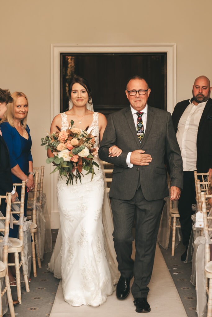 A bride in a white lace gown holding a bouquet walks down the aisle arm-in-arm with an older man in a grey suit. Guests seated on either side watch. The mood is joyful, and the room is softly lit with neutral decor and wooden chairs. © Aimee Lince Photography, Lincolnshire photographer