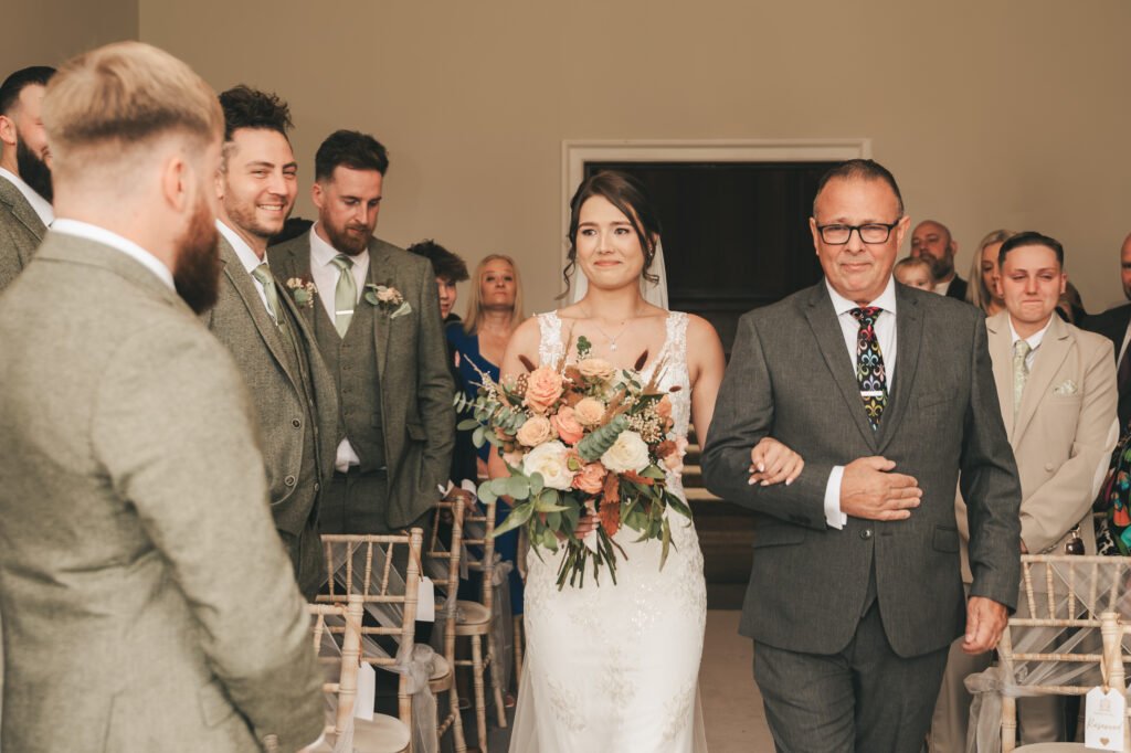 A bride in a white dress holds a large bouquet of flowers while walking down the aisle on her father’s arm. Groomsmen in grey suits stand to the left, smiling, as guests seated and standing watch the ceremony inside a softly lit room. © Aimee Lince Photography, Lincolnshire photographer