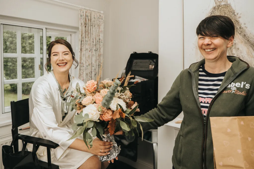Two women smiling indoors; one seated in a white dressing gown holding a large bouquet of pink, peach, and white flowers with greenery, the other standing in a striped shirt and green jacket holding a brown bag. A window and make-up case are visible in the bright room. © Aimee Lince Photography, Lincolnshire photographer