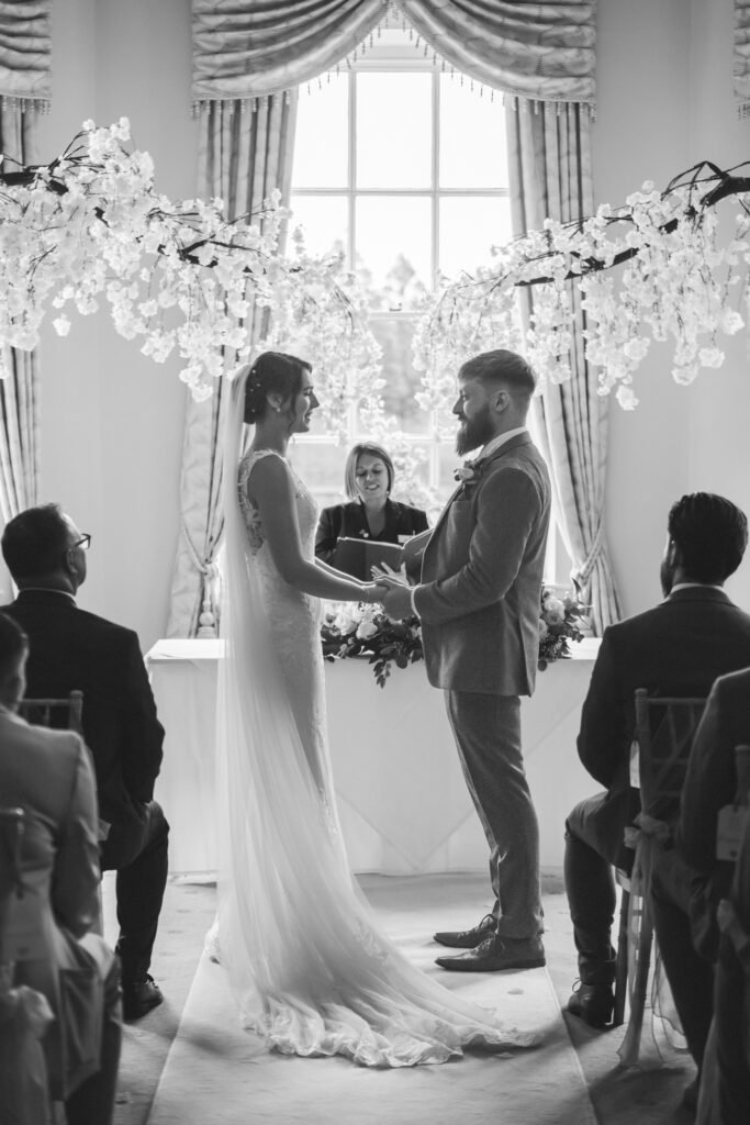 A bride and groom stand holding hands, facing each other under floral arches, during their wedding ceremony. An officiant stands behind a table, framed by tall windows and elegant curtains. Guests sit in rows, witnessing the moment. The image is in black and white. © Aimee Lince Photography, Lincolnshire photographer