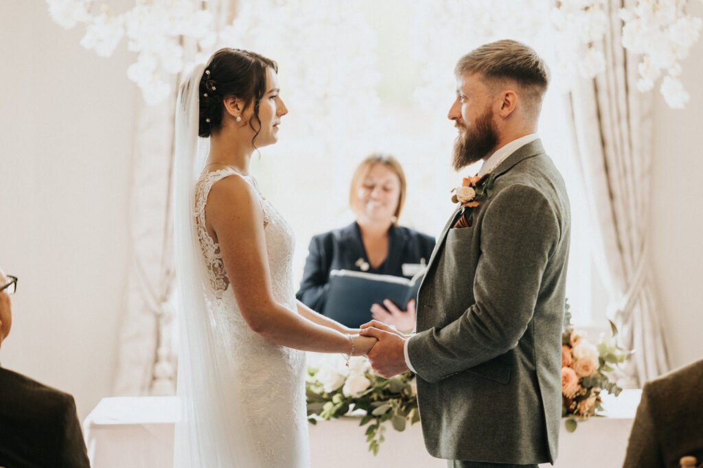 A bride and groom stand facing each other, holding hands and smiling during a wedding ceremony. The bride wears a white lace dress and veil, whilst the groom wears a light grey suit. An officiant with a book stands behind them. Floral decorations are in the background. © Aimee Lince Photography, Lincolnshire photographer