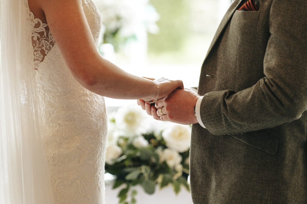 A bride in a lace wedding dress and veil holds hands with a groom in a grey suit with a pocket square. They face each other, with white flowers and greenery softly blurred in the background, suggesting a wedding ceremony. © Aimee Lince Photography, Lincolnshire photographer
