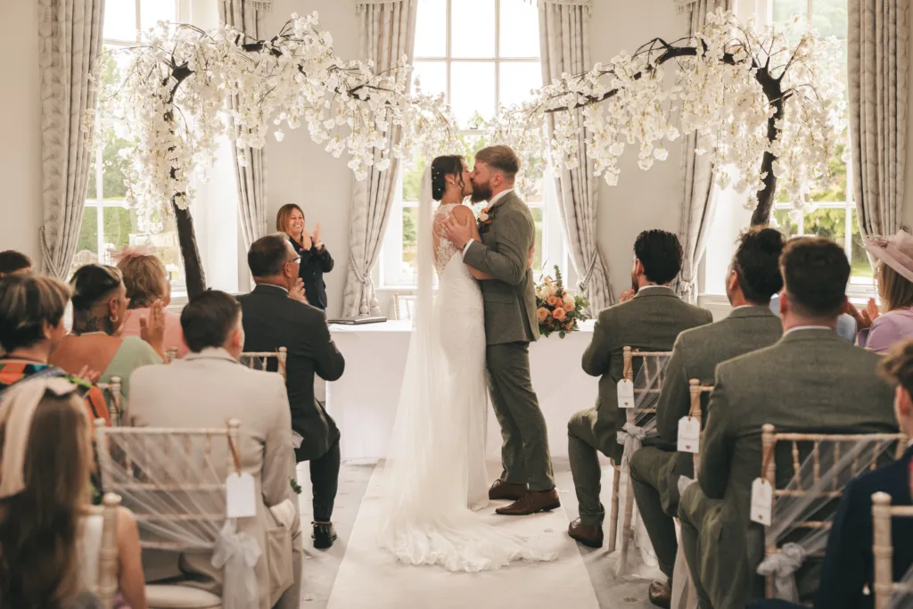 A bride and groom share a kiss under white floral arches in a sunlit room during their wedding ceremony, while guests seated on both sides watch. An officiant stands behind them at a white table, and large windows let in natural light. © Aimee Lince Photography, Lincolnshire photographer