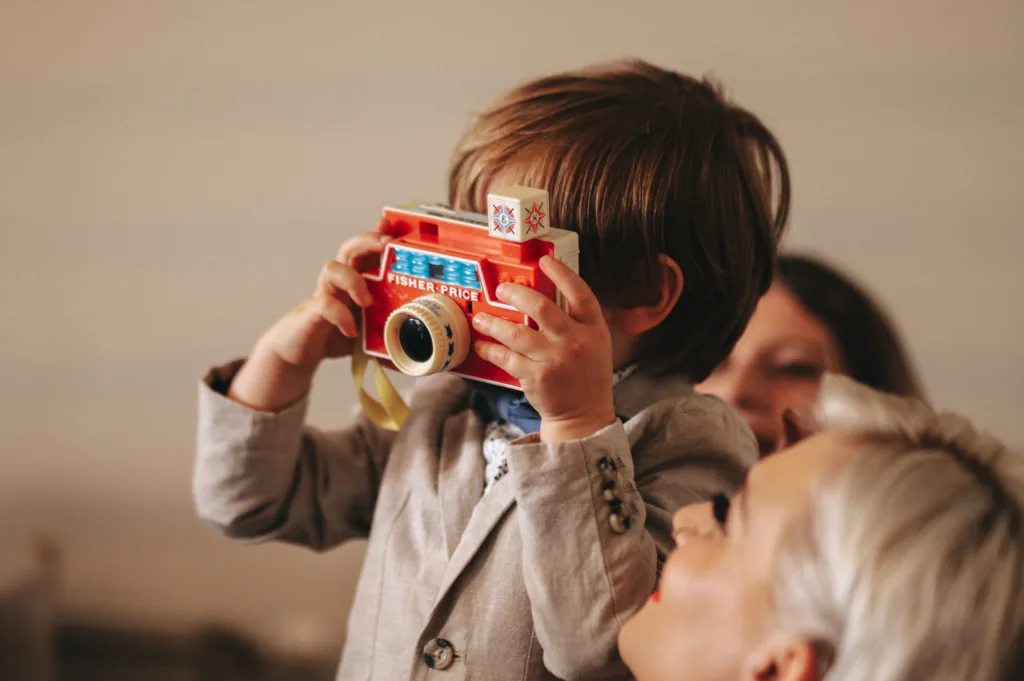 A young child in a grey suit holds a colourful Fisher-Price toy camera up to their face, pretending to take a photo. An adult with short, fair hair smiles in the foreground, and another person is slightly out of focus in the background. © Aimee Lince Photography, Lincolnshire photographer