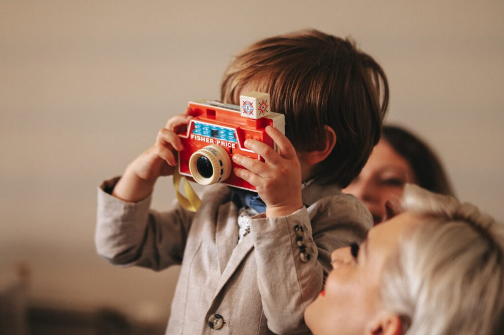 A young child in a grey suit holds a colourful Fisher-Price toy camera up to their face, pretending to take a photo. An adult with short, fair hair smiles in the foreground, and another person is slightly out of focus in the background. © Aimee Lince Photography, Lincolnshire photographer