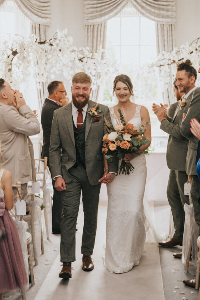A smiling bride in a white dress and veil holding a bouquet walks arm-in-arm with a bearded groom in a grey suit down the aisle. Guests on either side clap and smile. The room is decorated with white flowers and bright natural light floods in from tall windows. © Aimee Lince Photography, Lincolnshire photographer