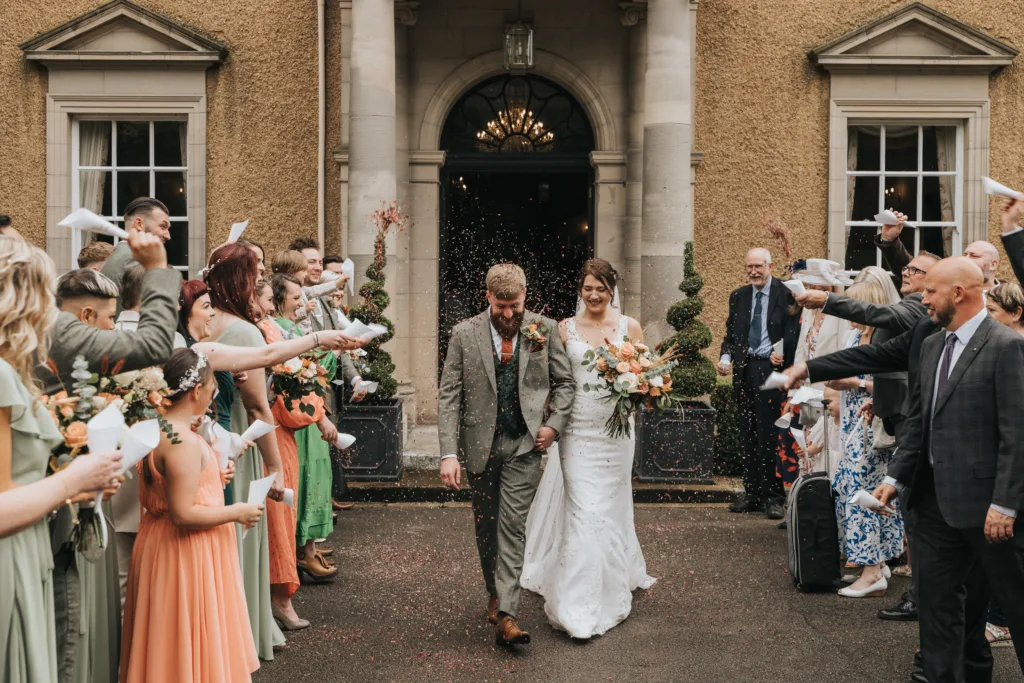 A newlywed couple smile as they walk hand-in-hand outside a building, surrounded by guests who throw confetti. The bride holds a bouquet and wears a white lace gown; the groom wears a tan suit. Guests in colourful attire stand on both sides, celebrating joyfully. © Aimee Lince Photography, Lincolnshire photographer
