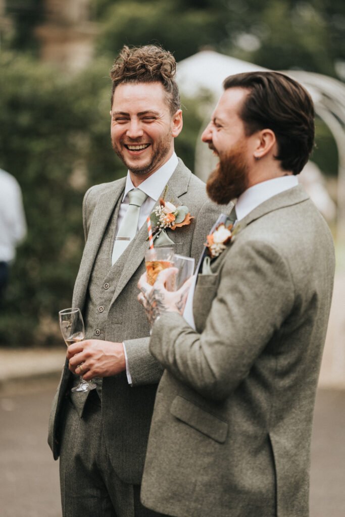 Two men in grey suits and pastel ties stand outdoors, smiling and laughing together. Each has a floral buttonhole. One holds a champagne glass, the other a cocktail with a striped straw. Green blurred foliage is in the background. © Aimee Lince Photography, Lincolnshire photographer