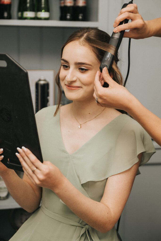 A young woman in a pale green dress smiles whilst looking into a black handheld mirror. Another person styles her hair with a curling tong. Shelves with bottles and a spray can are visible in the background. © Aimee Lince Photography, Lincolnshire photographer