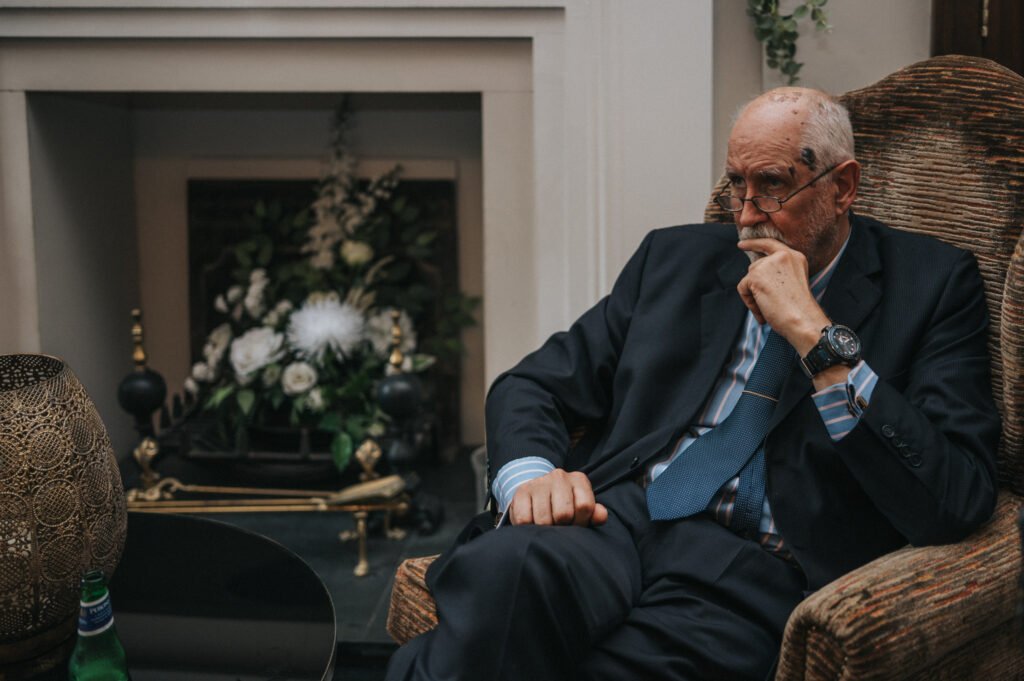 An older man in a suit and blue tie sits pensively in a brown armchair. He has glasses, a bald head, and rests his hand on his chin. Behind him is a fireplace with a floral arrangement. A table with a green bottle is in the foreground. © Aimee Lince Photography, Lincolnshire photographer