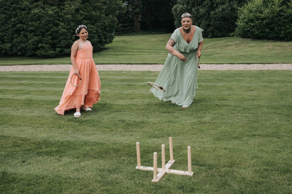 Two women in flowing dresses, one peach and one green, play a ring toss lawn game on grass. The woman in green throws a ring towards wooden pegs while the woman in peach smiles and watches. Lush greenery and hedges form the background. © Aimee Lince Photography, Lincolnshire photographer