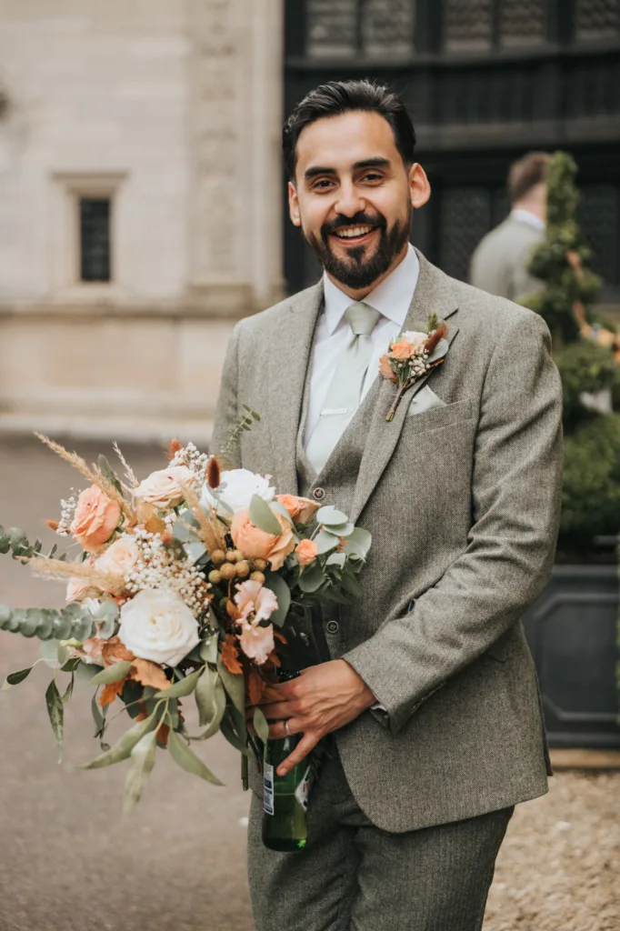A smiling man with dark hair and beard in a light green suit holds a large bouquet of peach and white flowers outdoors. He stands in front of an ornate stone building and greenery, wearing a buttonhole matching the bouquet. © Aimee Lince Photography, Lincolnshire photographer