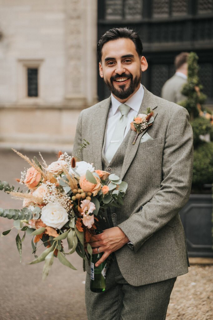 A smiling man with dark hair and beard in a light green suit holds a large bouquet of peach and white flowers outdoors. He stands in front of an ornate stone building and greenery, wearing a buttonhole matching the bouquet. © Aimee Lince Photography, Lincolnshire photographer