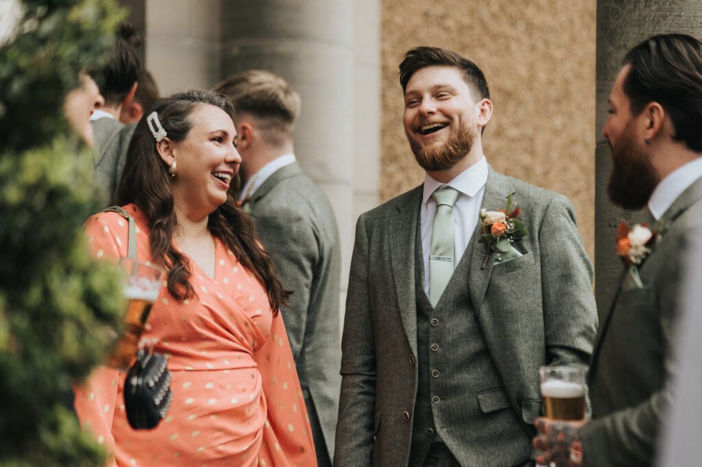 A woman in a coral polka-dot dress laughs with two men in grey suits holding beer glasses at an outdoor event. All are smiling and engaged in conversation, with greenery and stone columns in the background. © Aimee Lince Photography, Lincolnshire photographer