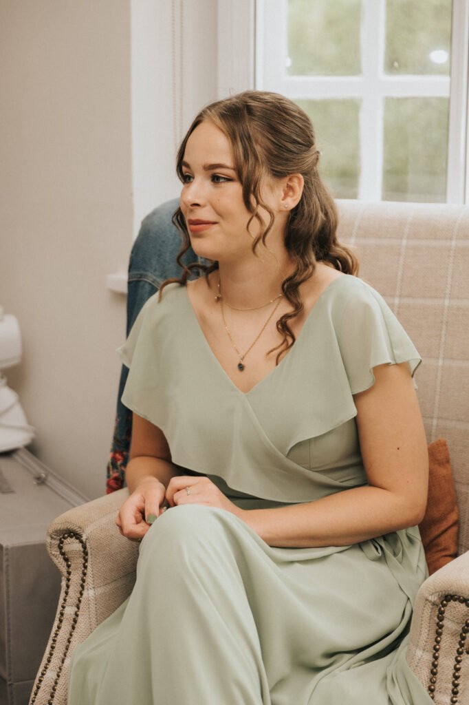 A young woman with wavy brown hair, wearing a light green, flutter-sleeve dress, sits on a beige armchair. She looks slightly to the side, smiling softly, with hands resting in her lap. A window and light-coloured walls appear in the background. © Aimee Lince Photography, Lincolnshire photographer