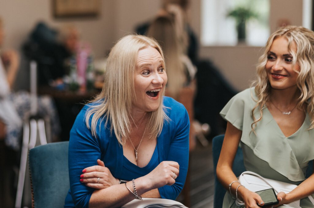 Two women sit indoors at a social event. The woman on the left, with straight blonde hair and a blue top, has an open-mouthed, joyful expression. The woman on the right, also blonde, wears a light green dress and smiles softly. Blurred people and decor are in the background. © Aimee Lince Photography, Lincolnshire photographer