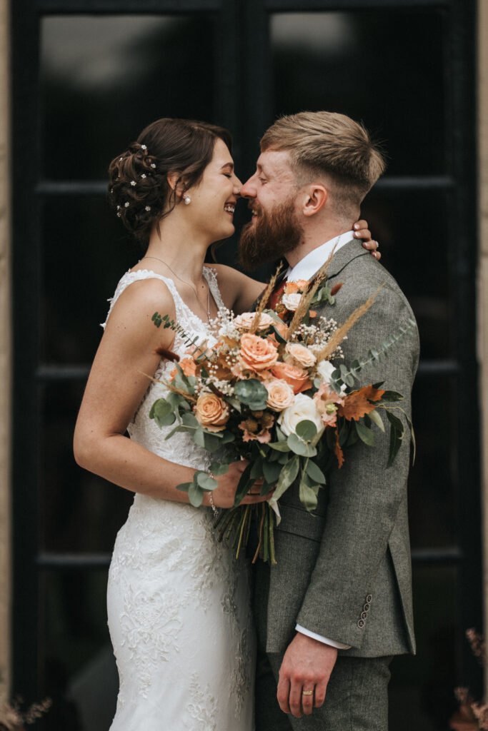 A smiling bride in a white lace dress holds a bouquet of peach and white roses with greenery, embracing a bearded groom in a grey suit and white shirt. They stand close together, touching noses, against a dark window background. © Aimee Lince Photography, Lincolnshire photographer