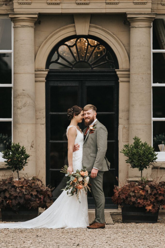 A bride in a white lace gown holds a bouquet and embraces a groom in a grey suit. They stand smiling in front of a grand building with arched windows, trimmed bushes, and beige stone columns, creating a romantic wedding scene. © Aimee Lince Photography, Lincolnshire photographer
