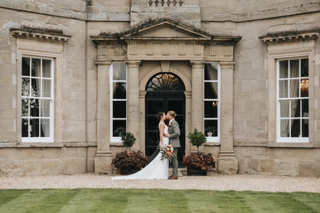 A bride in a white dress and a groom in a grey suit embrace and gaze at each other, standing in front of Bourton Hall in Warwickshire a grand stone building with large windows and a decorative doorway. The bride holds a bouquet, and green grass is in the foreground. © Aimee Lince Photography, Lincolnshire photographer