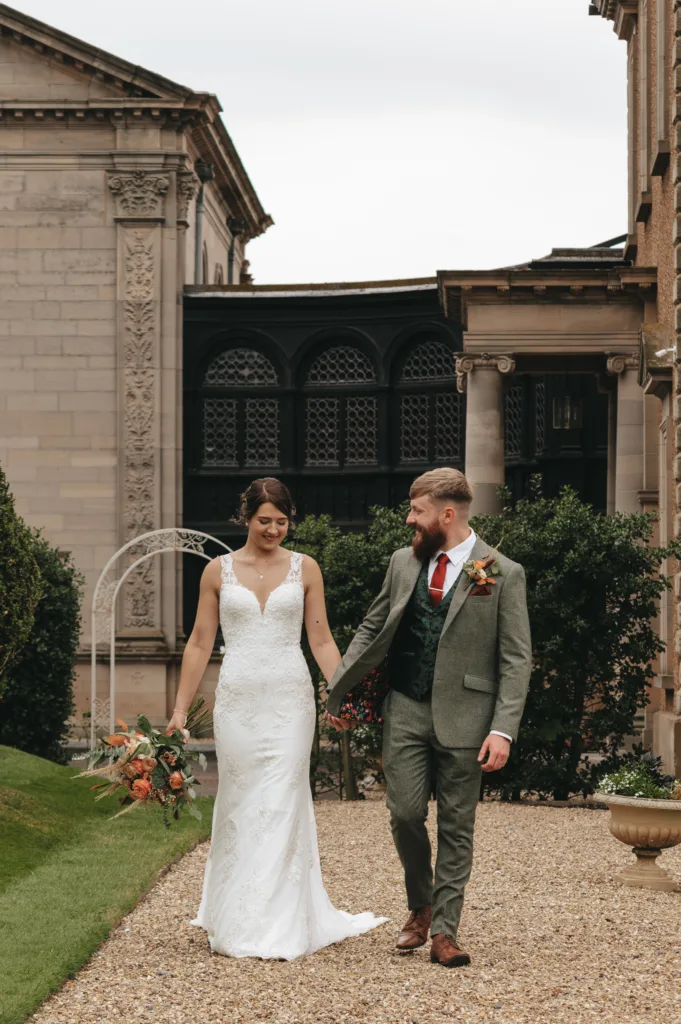 A bride in a white lace gown holding a bouquet walks hand-in-hand with a groom in a green suit, red tie, and buttonhole. They smile at each other outdoors, surrounded by greenery and elegant stone buildings with ornate architectural details. © Aimee Lince Photography, Lincolnshire photographer