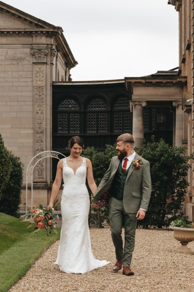 A bride in a white lace gown holding a bouquet walks hand-in-hand with a groom in a green suit, red tie, and buttonhole. They smile at each other outdoors, surrounded by greenery and elegant stone buildings with ornate architectural details. © Aimee Lince Photography, Lincolnshire photographer
