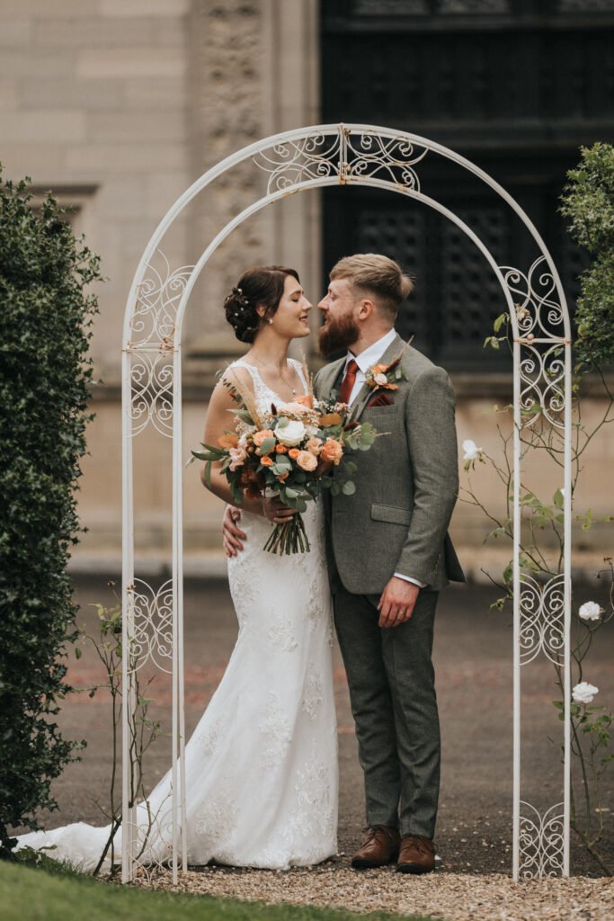 A bride in a white lace gown and a groom in a grey suit stand under a decorative white arch outdoors, smiling at each other. The bride holds a bouquet of orange and white flowers with greenery. The background features greenery and a stone building. © Aimee Lince Photography, Lincolnshire photographer