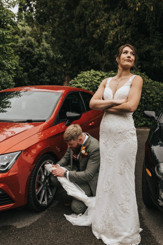 A bride in a white lace dress stands with arms folded, looking up and smiling, while a groom in a suit kneels beside her, cleaning her shoe with her dress near the tyre of a red car. Lush greenery and another black car are in the background. © Aimee Lince Photography, Lincolnshire photographer