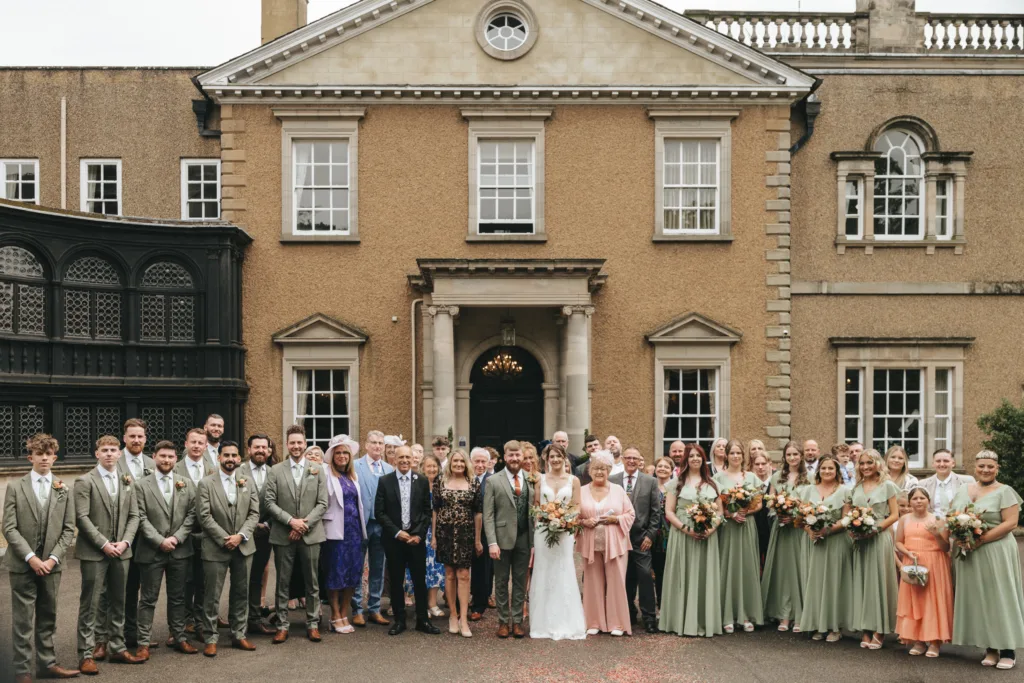 A large wedding group poses in front of Bourton Hall in Warwickshire, a grand, tan historic building. The bride and groom stand centre, surrounded by bridesmaids in sage green dresses and groomsmen in light suits. Family and friends in formal attire flank the couple, all smiling. © Aimee Lince Photography, Lincolnshire photographer