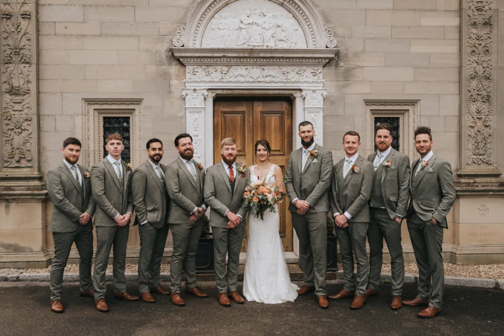 A bride in a white dress holds a bouquet and stands surrounded by ten groomsmen in matching grey suits, white shirts, and brown shoes, posing in front of an ornate stone building with carved pillars and a large double door. © Aimee Lince Photography, Lincolnshire photographer