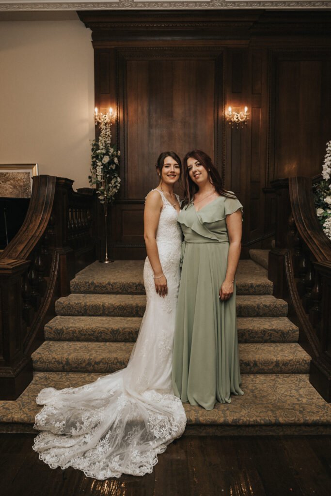 Two women stand side by side on carpeted stairs. One wears a white lace wedding dress with a train, the other wears a sage green gown. Both smile at the camera, with wood-panelled walls, floral arrangements, and warm lighting in the background. © Aimee Lince Photography, Lincolnshire photographer