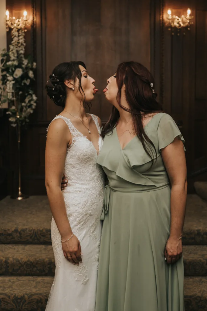 Two women stand close, facing each other and sticking out their tongues playfully. One wears a white lace wedding dress; the other wears a sage green gown. They stand on carpeted steps in front of dark wooden doors with floral arrangements and wall sconces behind them. © Aimee Lince Photography, Lincolnshire photographer