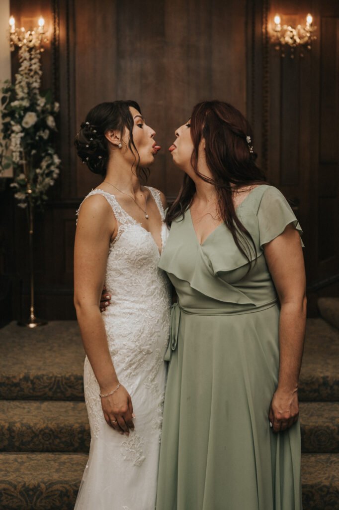 Two women stand close, facing each other and sticking out their tongues playfully. One wears a white lace wedding dress; the other wears a sage green gown. They stand on carpeted steps in front of dark wooden doors with floral arrangements and wall sconces behind them. © Aimee Lince Photography, Lincolnshire photographer