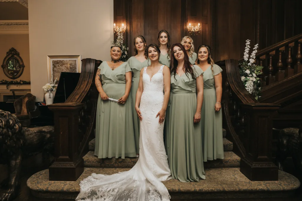 A bride in a white gown stands on a staircase flanked by six bridesmaids in sage green dresses. They all face forward, smiling. The background features dark wood panelling, two wall sconces, and floral arrangements on each side of the staircase. © Aimee Lince Photography, Lincolnshire photographer